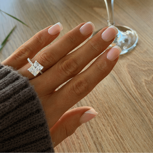 A close-up of a hand wearing a silver ring with a large clear rectangular gemstone, resting on a wooden table with a cocktail glass in the background.