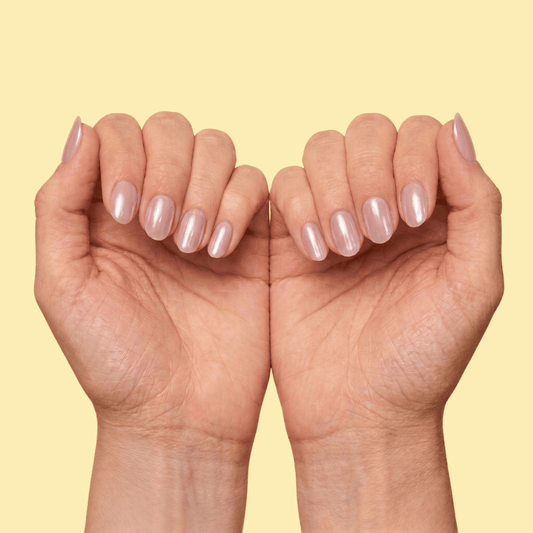 A close-up of two hands with short, oval-shaped nails painted in a glossy, pale pinkish-beige shade, set against a solid pale yellow background.