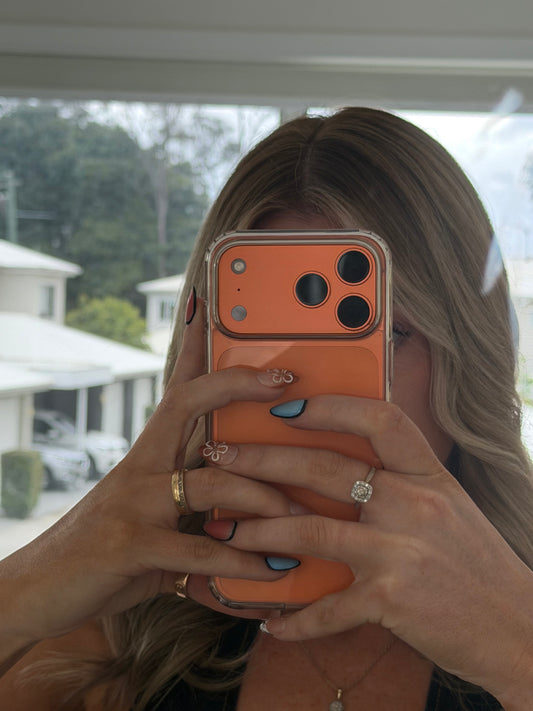 A woman taking a mirror selfie with her hands holding a phone with a bright orange protective case. She has blonde hair, blue nail polish, and is wearing rings.