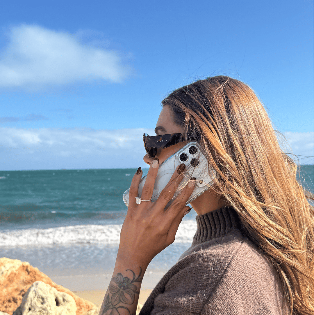 A woman with long light brown hair wearing a brown knit turtleneck sweater and sunglasses is shown in profile, holding a white smartphone to her ear outdoors by the ocean with rocks and blue sky in the background.