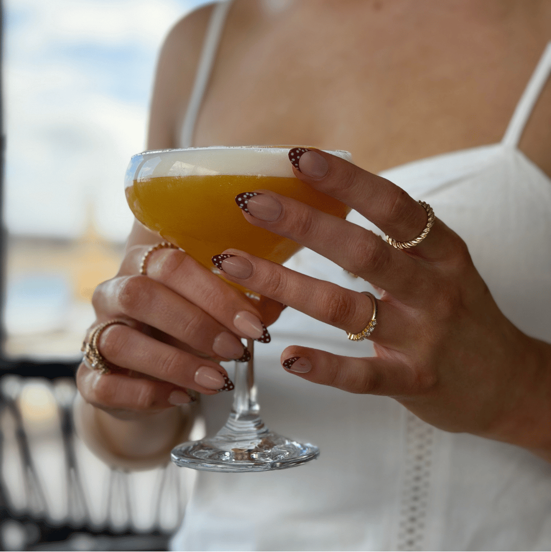 A close-up lifestyle shot of a woman holding a cocktail in a coupe glass, wearing a white spaghetti-strap top and multiple gold rings, with a blurred beach background.