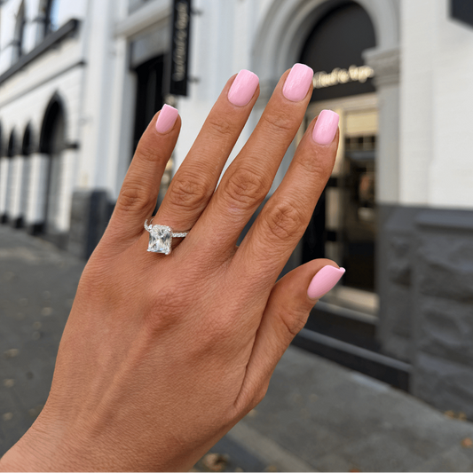 A close-up of a hand with light pink manicured nails wearing a silver-toned ring with a large clear gemstone, set against a blurred city street background.