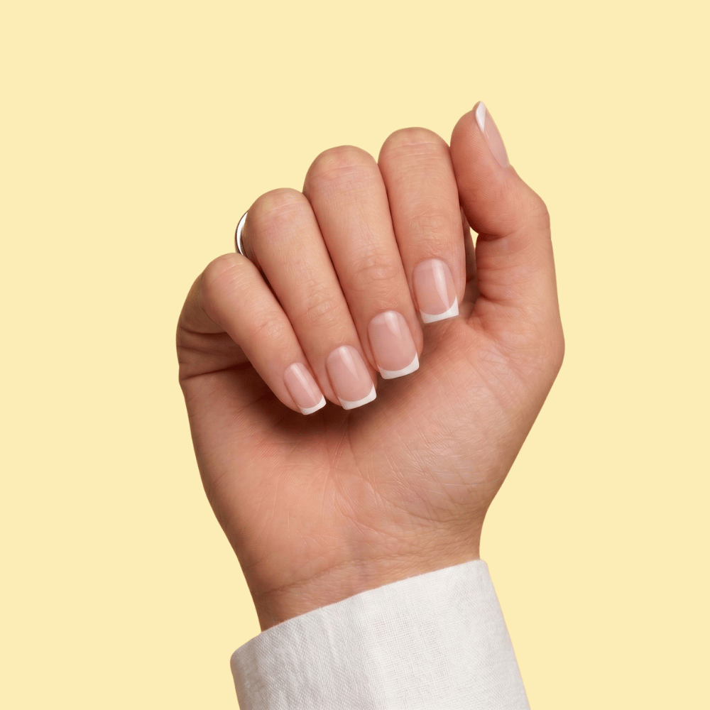 A close-up of a hand with a classic French manicure, featuring a white tip and a soft nude-pink base, set against a solid pale yellow background.