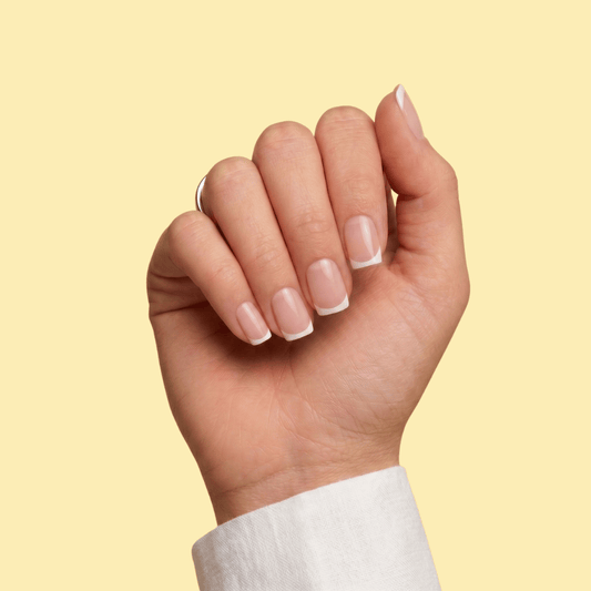 A close-up of a hand with a classic French manicure, featuring a white tip and a soft nude-pink base, set against a solid pale yellow background.