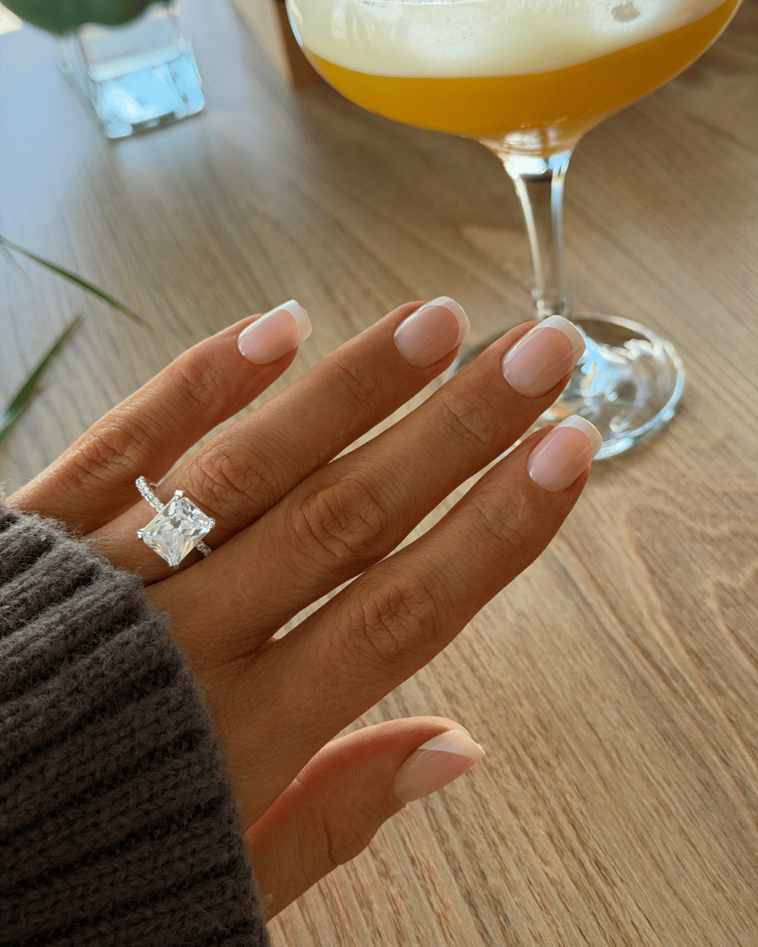 A close-up of a hand wearing a silver ring with a large clear rectangular gemstone, resting on a wooden table with a cocktail glass in the background.