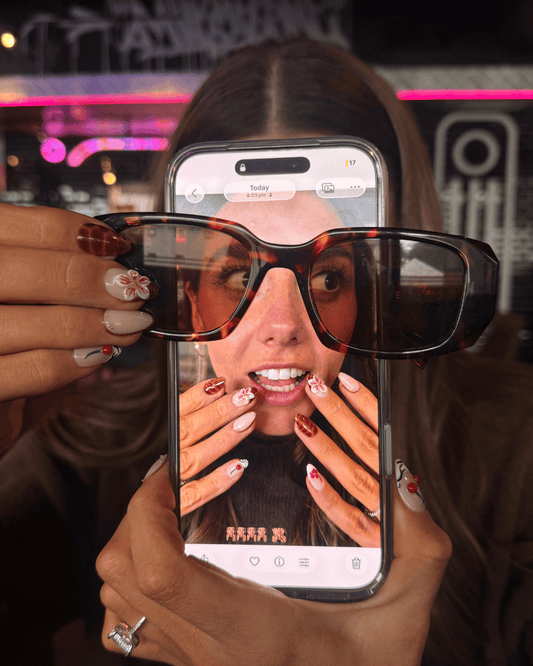 A person holds a smartphone in front of their face, using the screen to demonstrate a social media-style filter or editing effect. The phone is positioned behind a pair of tortoiseshell eyeglasses, and the visible image on the screen shows a distorted, stylized version of the person's face and hands.