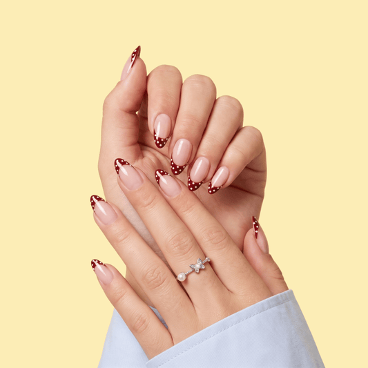 A close-up of two hands with manicured nails featuring a glossy nude-pink base, red polka dots, and a small white star accent near the tips. One hand wears a silver ring with a clear stone.