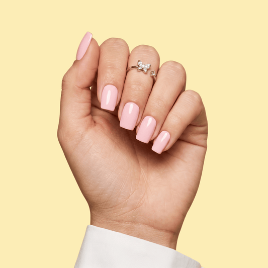 A close-up of a hand with glossy light pink manicured nails, wearing a small silver ring, against a solid pale yellow background.