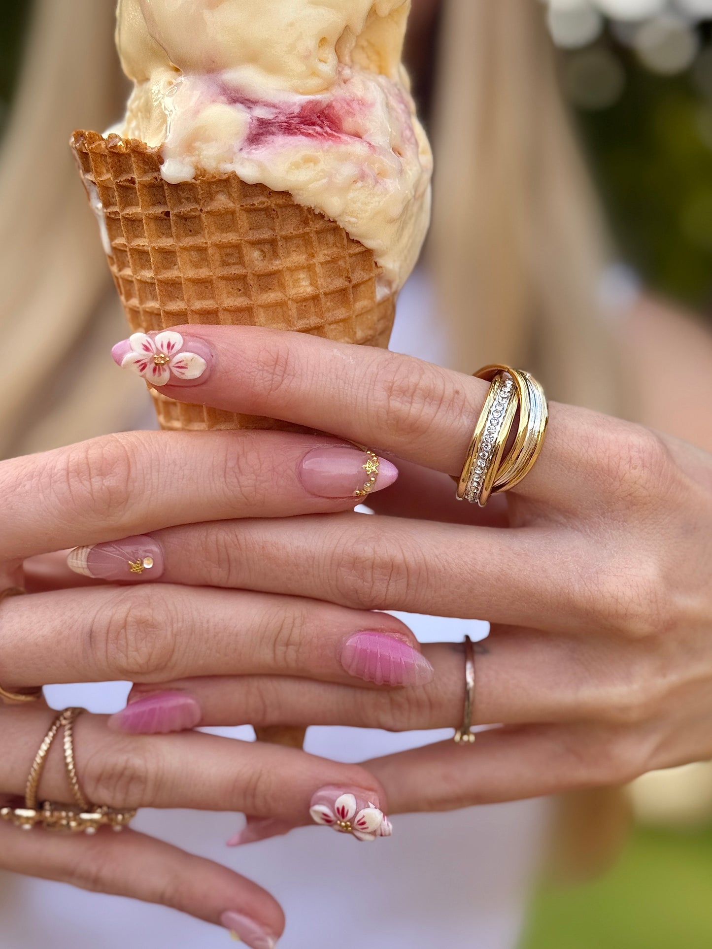 Close-up of hands holding an ice cream cone, wearing multiple gold rings including one with small clear stones and floral nail art with pink and white accents.