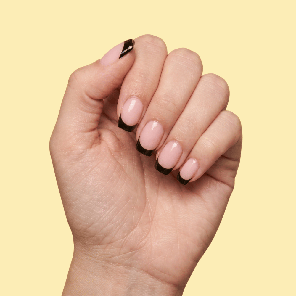 A close-up of a hand with a classic French manicure, featuring light pink nails with black tips, set against a solid pale yellow background.