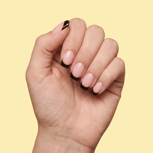 A close-up of a hand with a classic French manicure, featuring light pink nails with black tips, set against a solid pale yellow background.