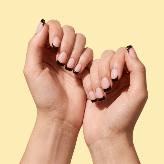 Close-up of two hands with manicured nails featuring a pale pink base and black French tips, posed against a solid pale yellow background.