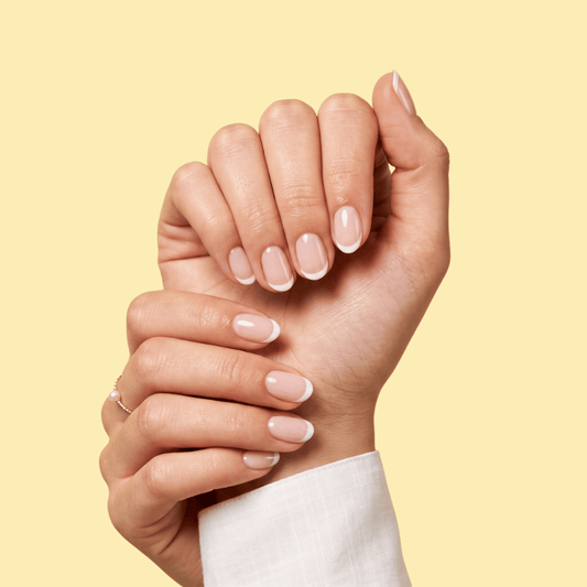A close-up of two hands with a classic French manicure, showcasing long, oval-shaped nails with a white tip and nude pink base, set against a solid pale yellow background.