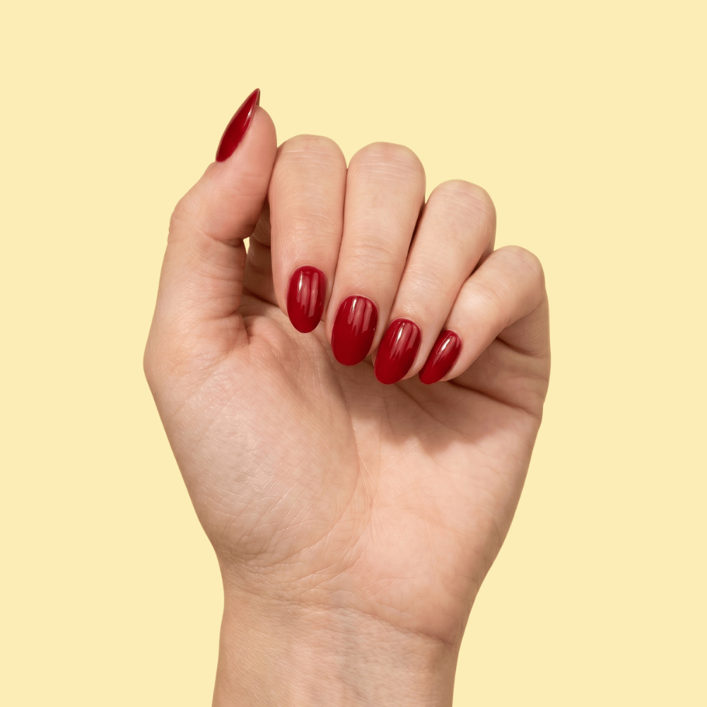 A close-up of a hand with glossy, almond-shaped nails painted in a deep red shade, set against a solid pale yellow background.
