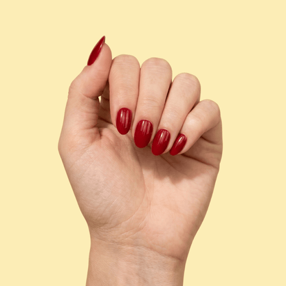 A close-up of a hand with glossy, almond-shaped nails painted in a deep red shade, set against a solid pale yellow background.
