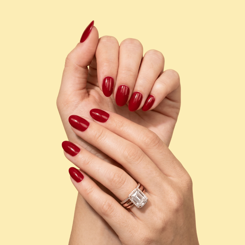 A close-up of two hands with glossy dark red manicured nails, one wearing a rose gold band with a large emerald-cut diamond ring, set against a solid pale yellow background.