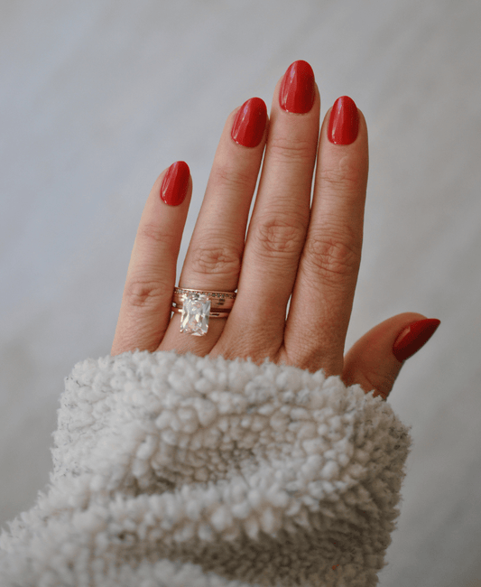 A close-up of a hand with red manicured nails wearing a gold ring with a clear rectangular gemstone, set against a soft gray background and a white fuzzy sleeve.