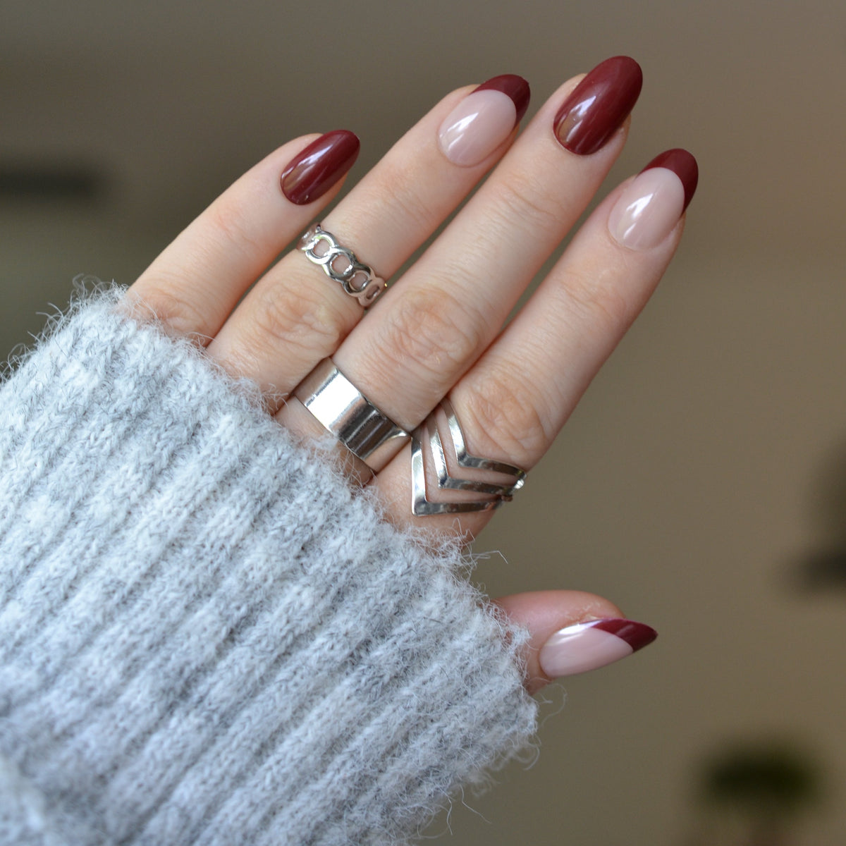 A close-up of a hand wearing multiple silver rings, including a wide band, a chevron-patterned ring, and a textured geometric ring, with manicured nails in a glossy dark red and nude pink color combination, set against a soft, out-of-focus neutral background.