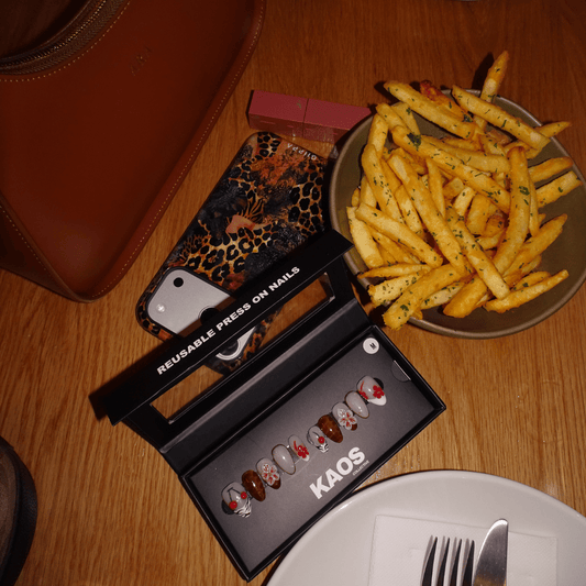 A top-down view of a styled table scene featuring a black KAOS reusable press-on nails set in its box, next to a plate of french fries, a leopard print phone case, and a brown leather bag on a wooden table.