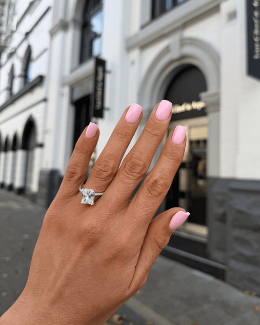 A close-up of a hand with light pink manicured nails wearing a silver-toned ring with a large clear gemstone, set against a blurred city street background.