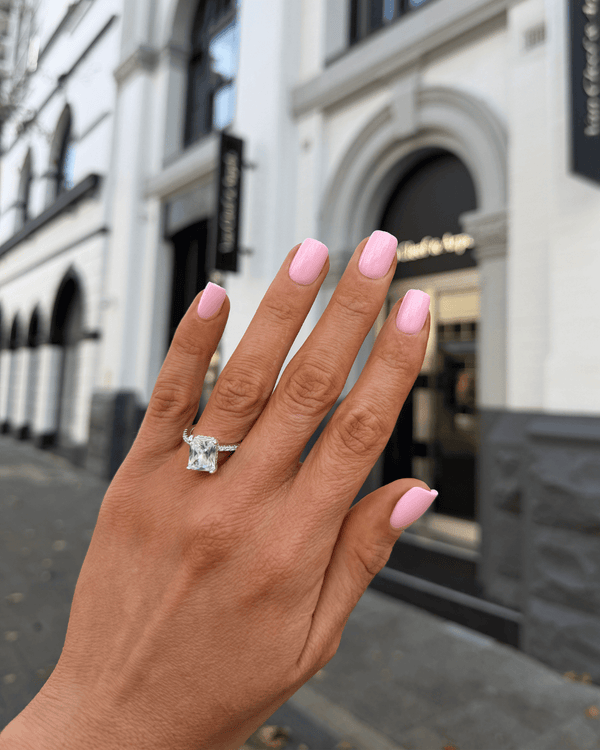 A close-up of a hand with light pink manicured nails wearing a silver-toned ring with a large clear gemstone, set against a blurred city street background.