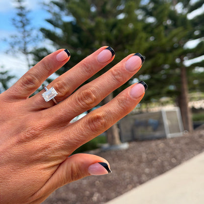 A close-up of a hand wearing a silver-toned ring with a square-cut clear gemstone, set against a blurred outdoor background of trees and sky.