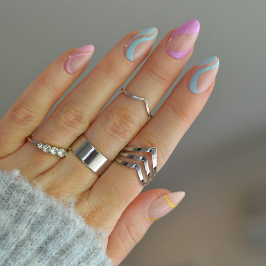 A close-up of a hand with pastel-colored manicured nails wearing multiple silver rings, including a wide silver band, a ring with small clear stones, and a geometric chevron-style ring. The hand is wearing a light gray knit sweater, set against a soft, blurred neutral background.